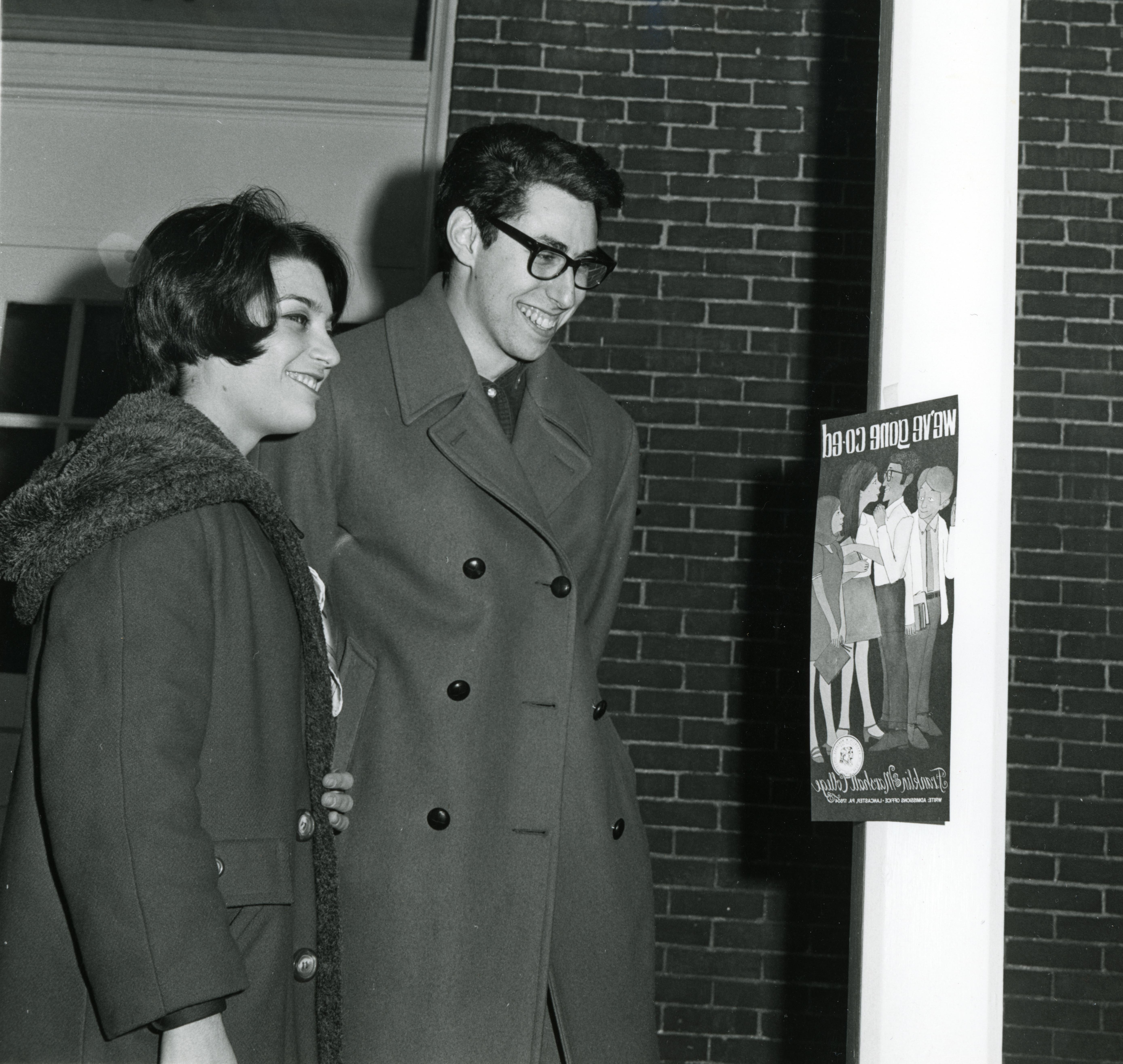 Two students looking at a poster reading "We've gone coed." Notes on the verso suggest that the photograph was printed in an F&M publication.
